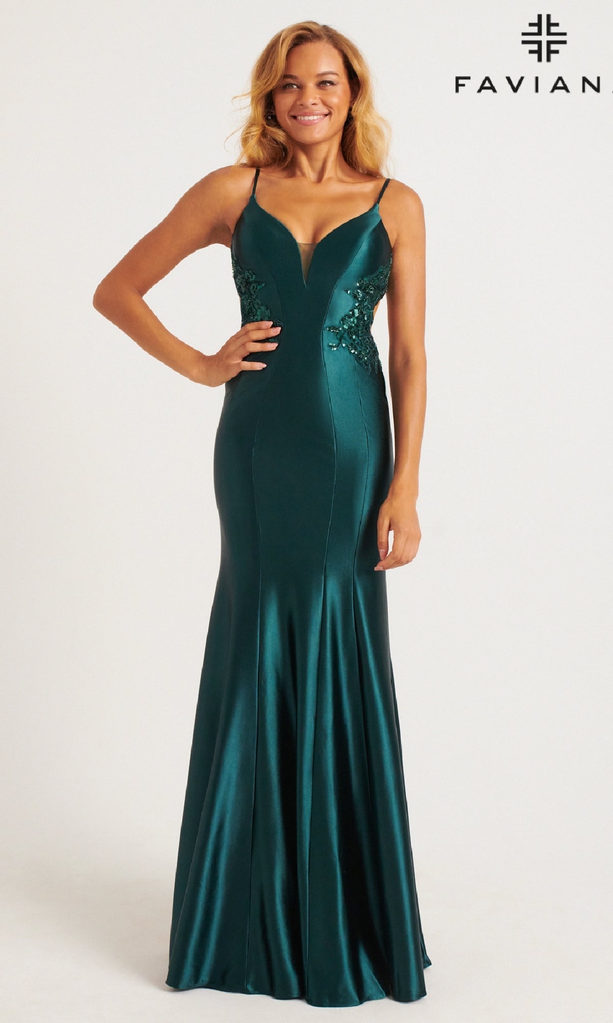 A woman stands against a plain white background wearing the Faviana Sheer-Lace Sides Long Prom Dress 11007, a sleeveless floor-length emerald green satin gown with lace detailing and a mermaid skirt.